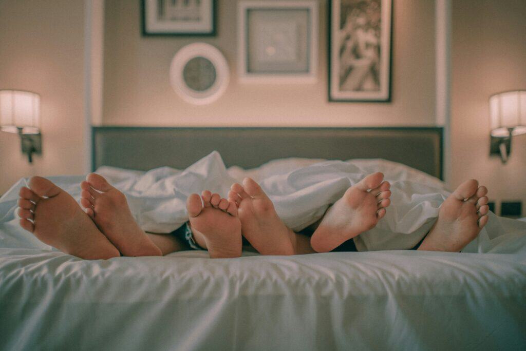 Sechs Füße die unten an der Bettdecke herausschauen lassen ahnen, dass drei Menschen im Bett liegen.