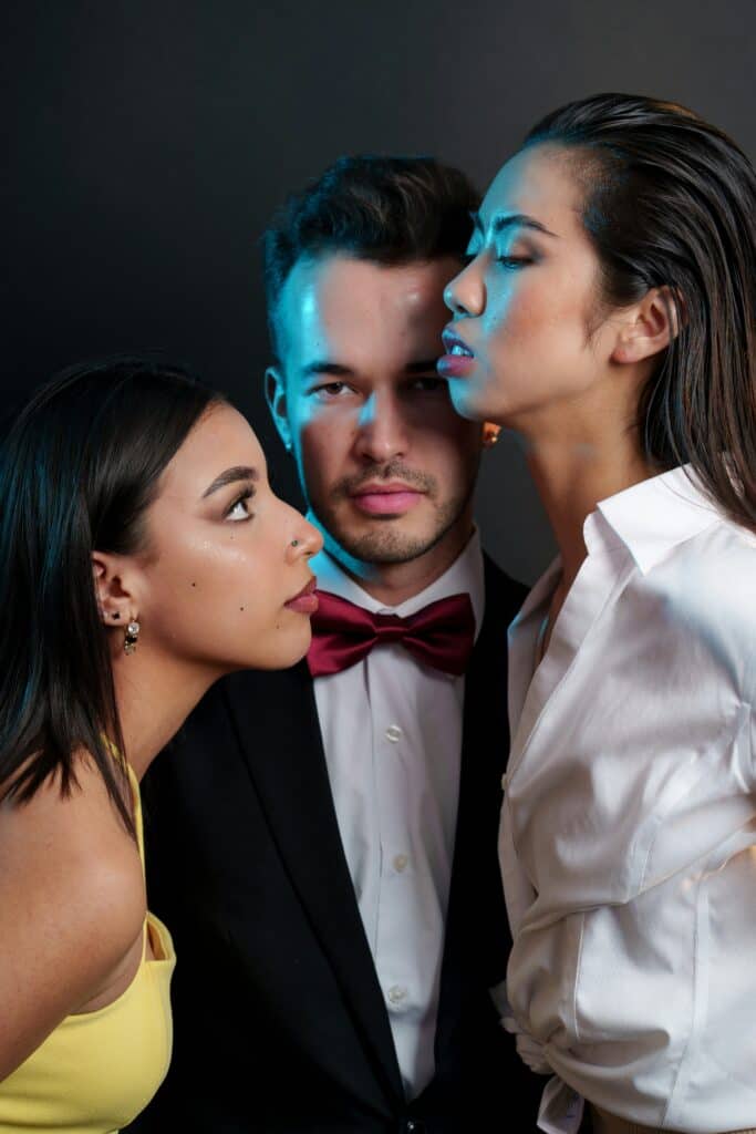 Elegant angezogene Personen mit einem Mann mit Smoking und Fliege in der Mitte stehend, dem zwei Frauen an seiner Seite ihre Aufmerksamkeit schenken.