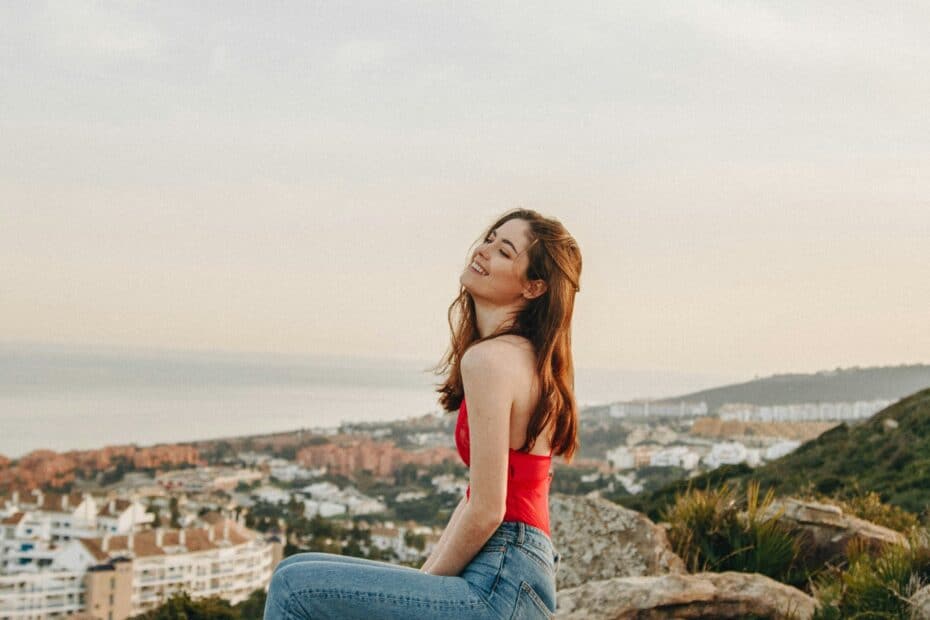 Coverbild zum Bericht über Diary Lady Escort mit einer jungen Frau mit roten Top und Jeans auf einem Hügel auf eine Stadt mit Strand schauend.